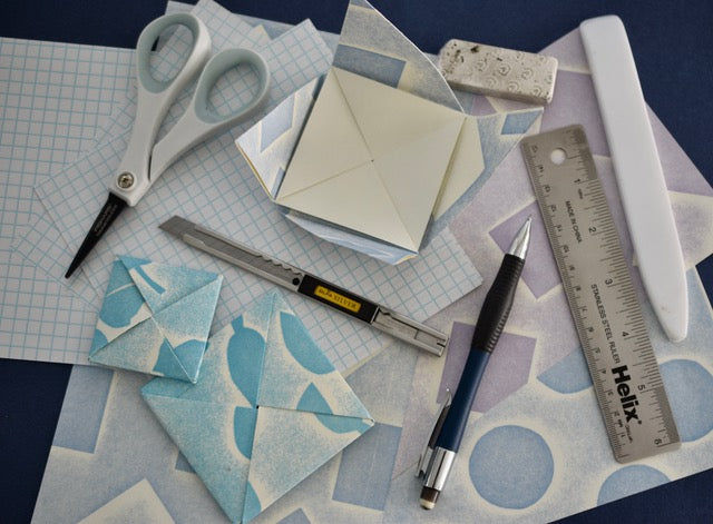 A flat-lay arrangement of origami crafting tools and finished paper models on patterned sheets, including scissors, a utility knife, a ruler, a pen, a small metal file, and folded blue and white origami pieces.