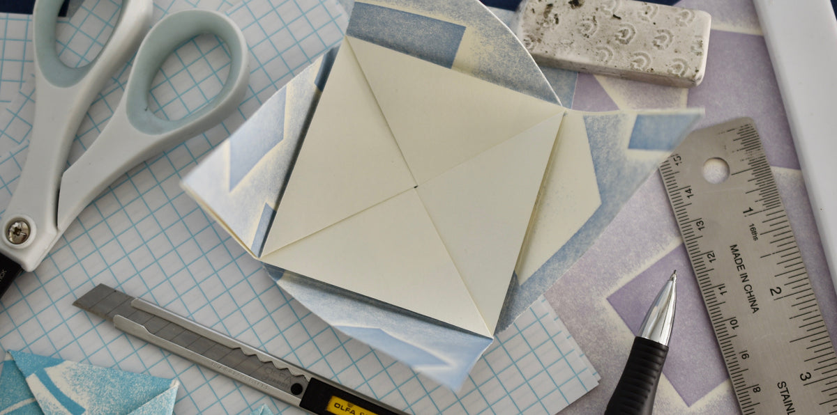 A flat-lay of crafting tools and materials, featuring a central cream-colored square paper envelope or pocket square, surrounded by scissors, a utility knife, a ruler, a pen, patterned paper, and a small metal weight on a dark blue background.